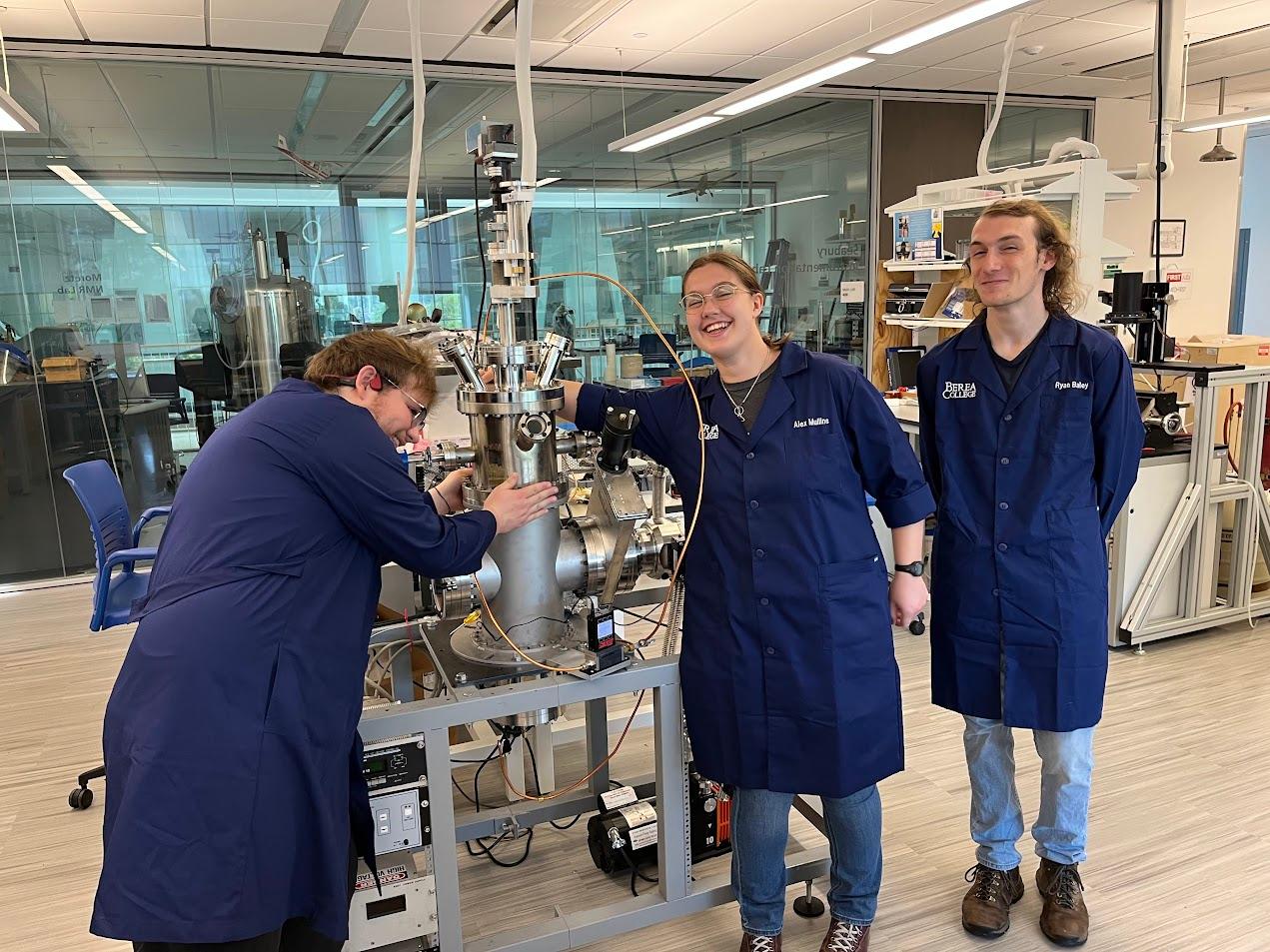 Three Berea College physics students smile at the camera as they work on a large machine in a lab classroom.