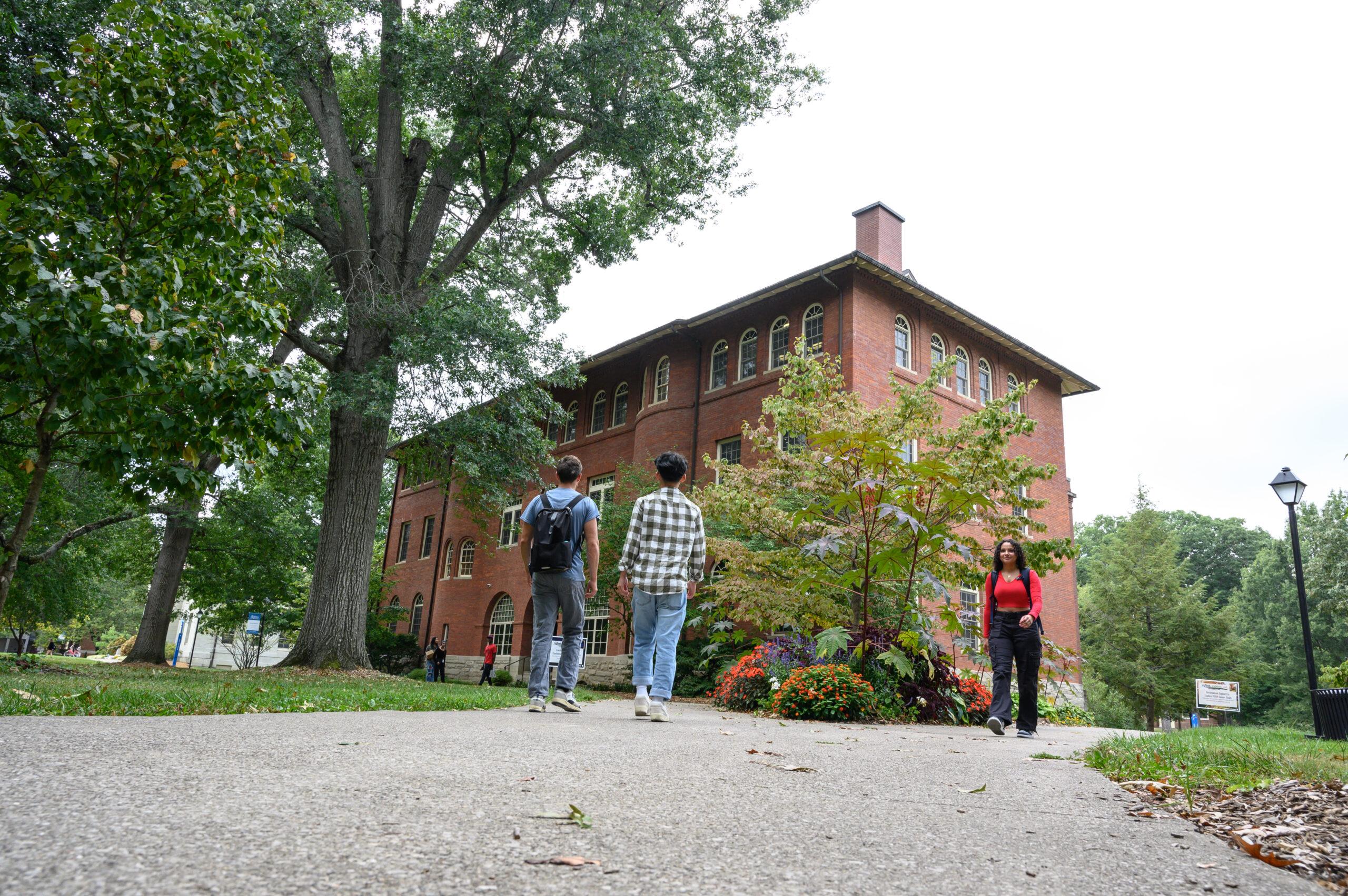 Photo of students walking on campus, two students walking together with their backs towards the camera on the left side of the photo, and another walking towards the camera on the right.